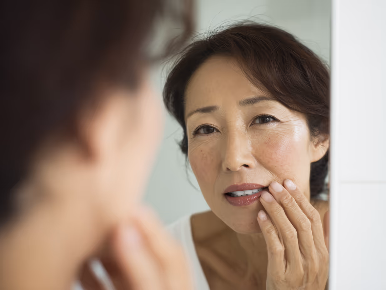 Middle-aged woman with short brown hair gently touching her cheek while looking in the mirror.