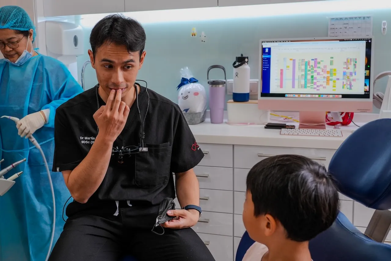 Dr Martin Nguyen in black scrubs demonstrating oral care to a child in OSD clinic with an assistant in blue protective clothing in the background.