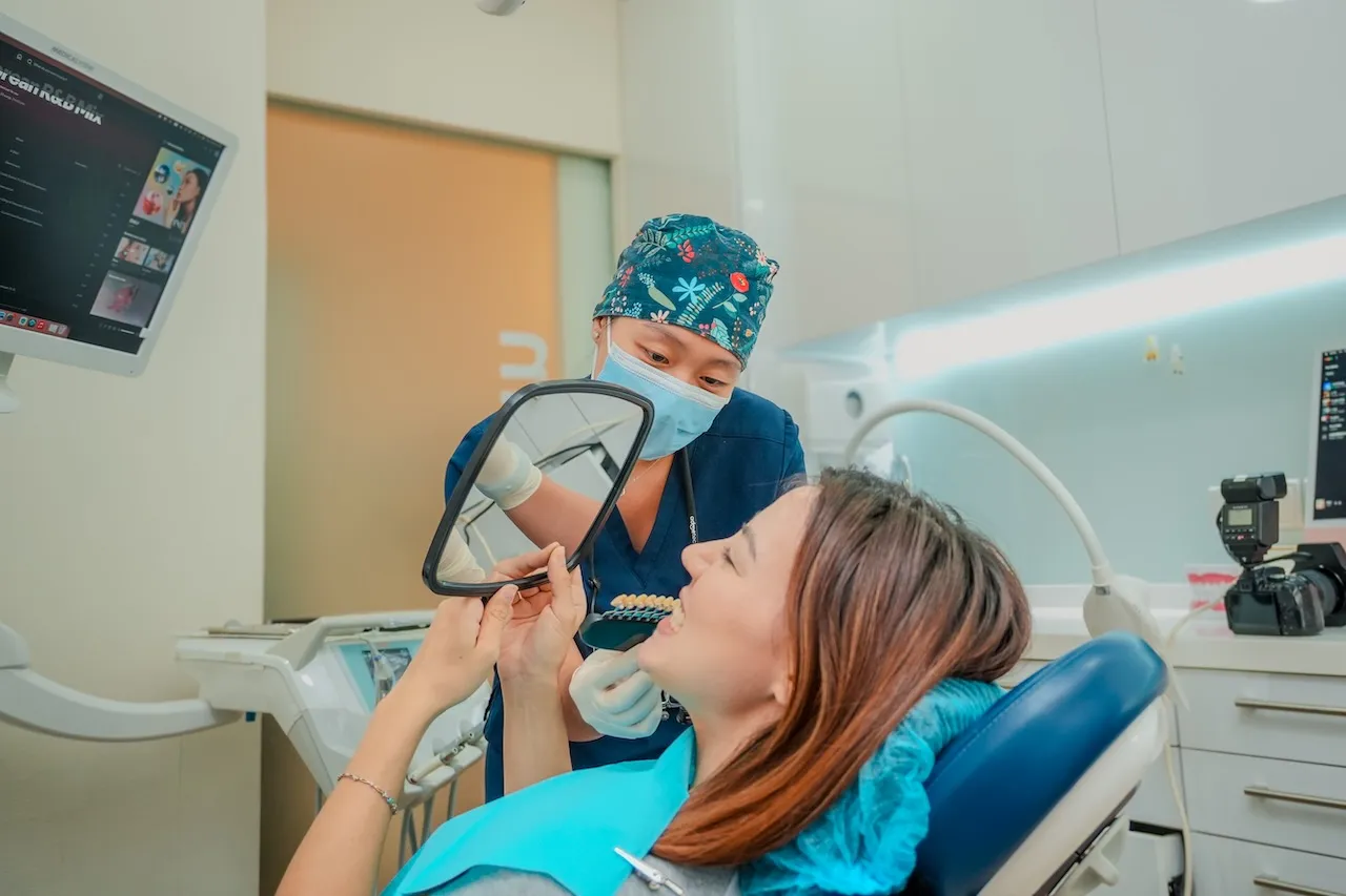 Dr Pearlyn Ng from Orchard Scotts Dental showing a smiling patient a mirror with a shade guide for teeth whitening in OSD clinic